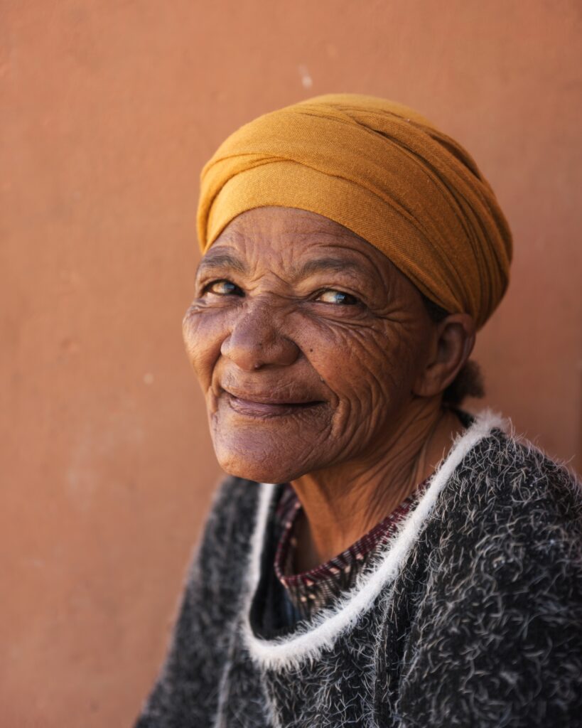 Portrait of an older woman wearing a mustard headscarf, smiling against a warm terracotta-coloured wall.