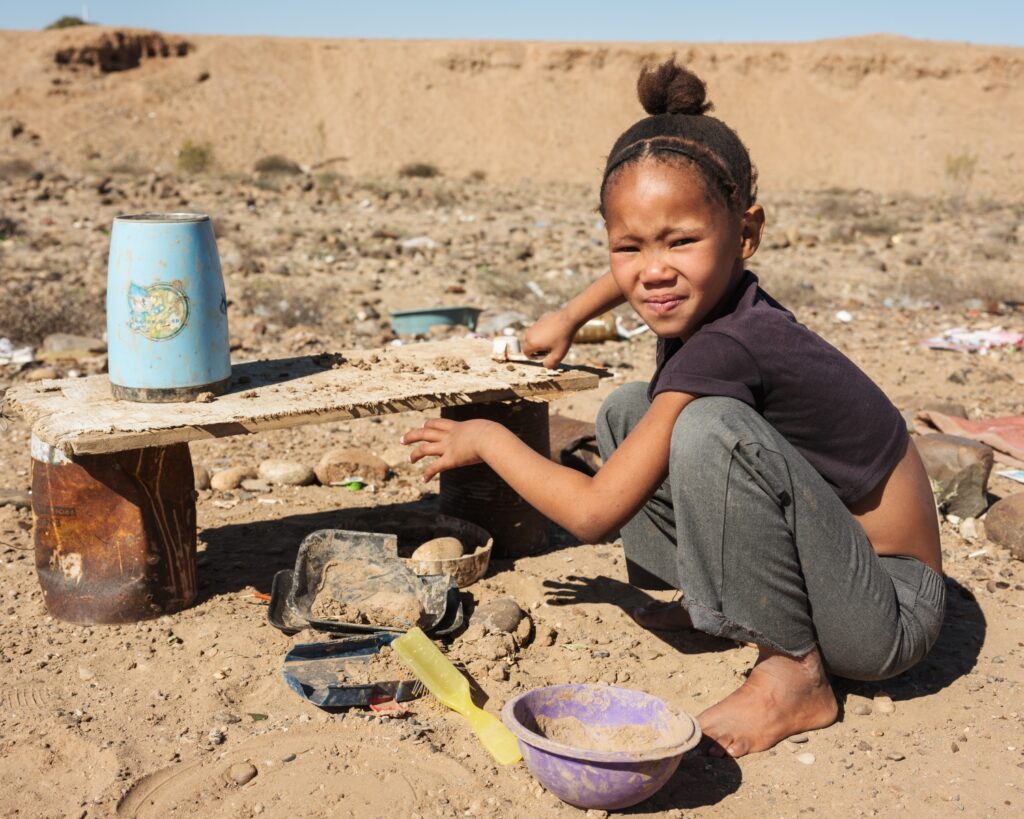 Young girl crouching barefoot on sandy ground beside bowls and household items in an arid outdoor setting.
