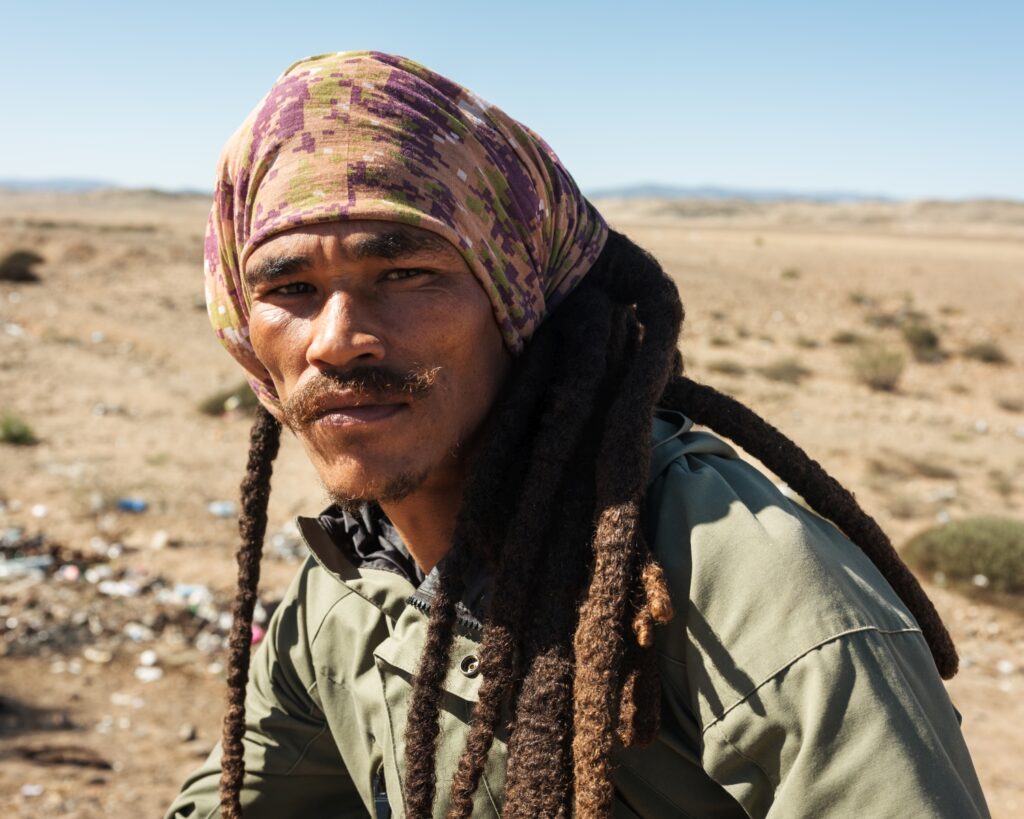 Portrait of a young man wearing a patterned headscarf, with an arid landscape in the background.