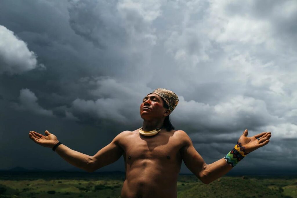 Indigenous man standing with arms outstretched beneath dramatic storm clouds in a remote natural landscape.