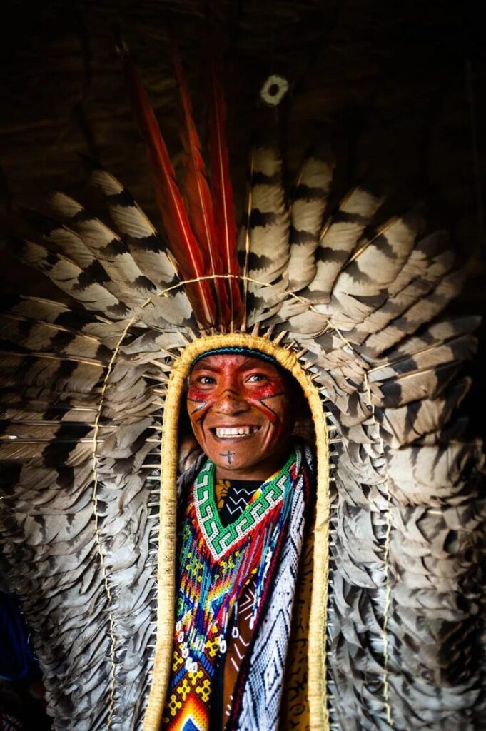 Smiling Indigenous man wearing an elaborate feather headdress, colourful beadwork and painted facial markings.