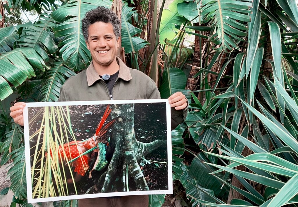Photographer Gary van Wyk smiling while holding a large print of one of his Amazon portraits, surrounded by lush tropical foliage.