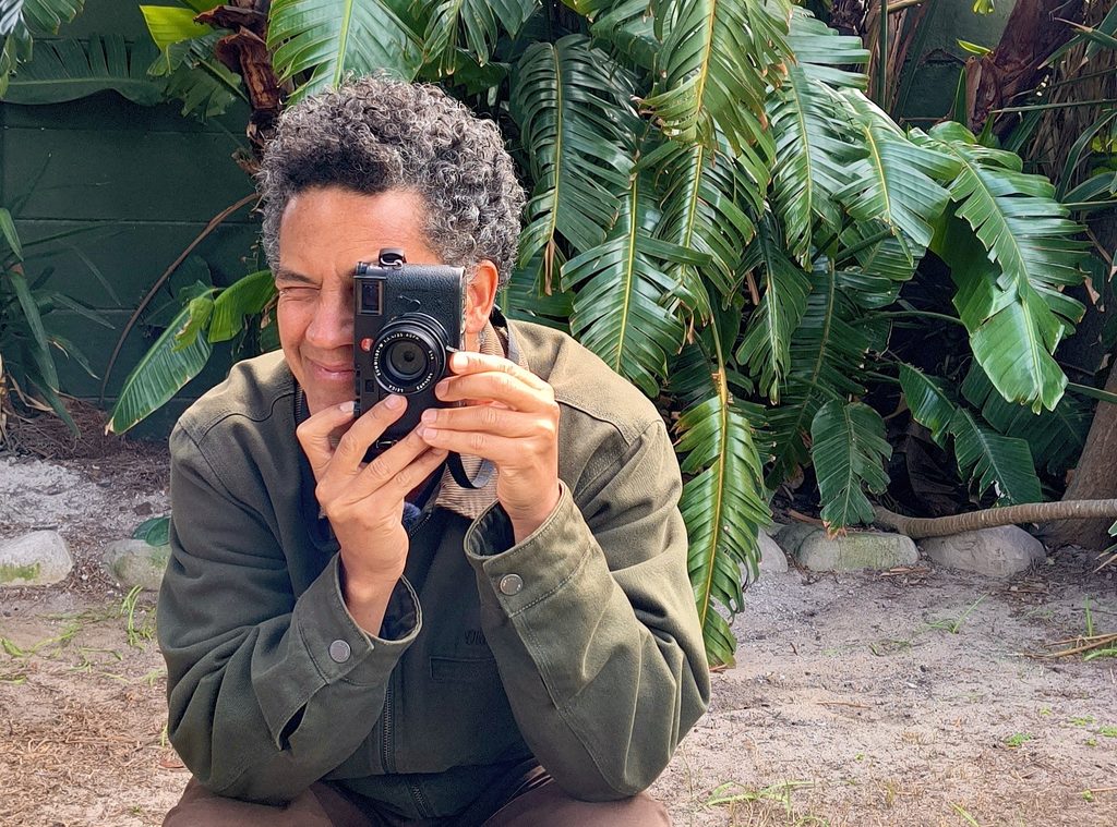Photographer Gary van Wyk smiling as he takes a photograph with a camera, with lush green foliage behind him.