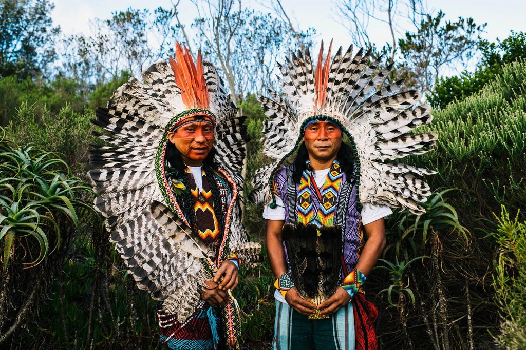 Two Indigenous men wearing elaborate feather headdresses and colourful traditional beadwork, standing together outdoors.
